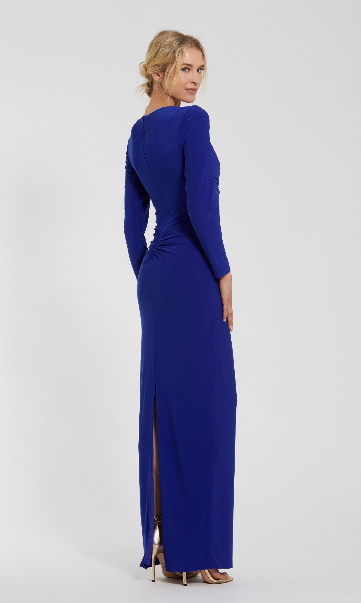 A woman, photographed from behind against a plain light backdrop, wears the Mac Duggal 44064 blue long sleeve formal dress with a back slit and pairs it with metallic high-heeled shoes.